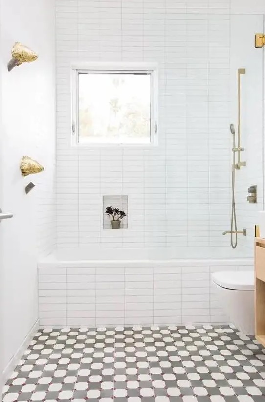 A bright bathroom with a mosaic floor, white tiles with built in shelves, a window and gold hardware