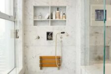 a chic shower space clad with white marble tiles, with niche with shelves used for storage, a folding seat, gold fixtures