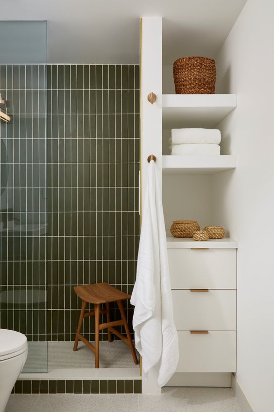A modern bathroom done in neutrals and dark green skinny tiles, a deep niche with shelves and a built in cabinet for storage