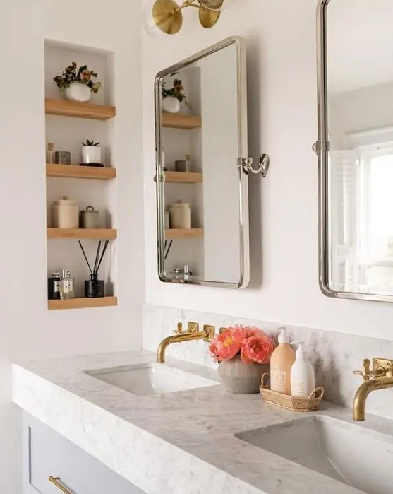 a modern bathroom with a double vanity and sinks, mirrors, a niche with wooden shelves and decor plus some blooms
