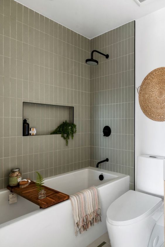 a modern bathroom with a tub and grey skinny tiles around it, a niche with a potted plant and some stuff and a woven piece on the wall