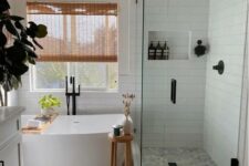a modern farmhouse bathroom clad with grey square and white skinny tiles, a shower space, an oval tub, a white vanity, wooden rugs and shades, a niche in the shower