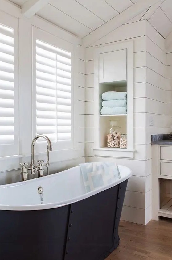 a modern farmhouse bathroom done with shiplap, with an oval tub, a window with shutters, a niche with shelves used for decor and towels