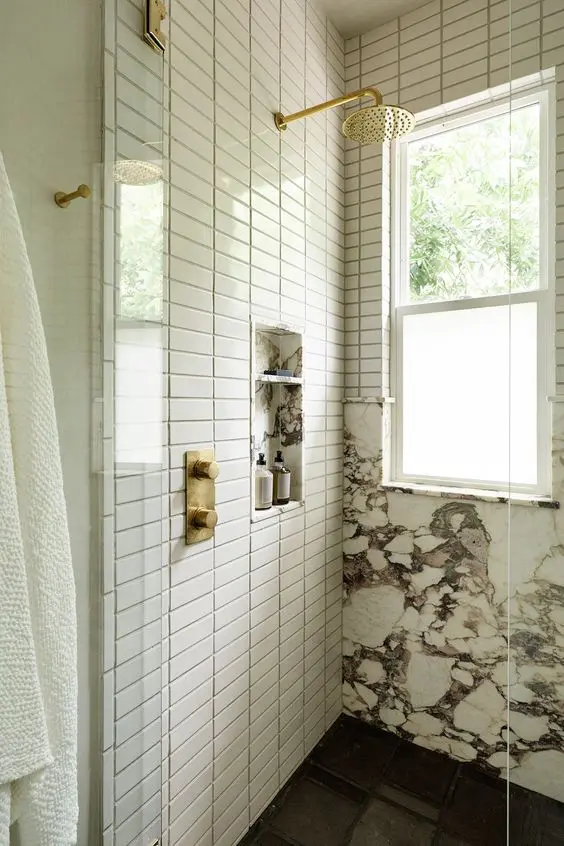 a modern shower space with marble and white skinny tiles, a niche for storage and gold fixtures is chic and refined