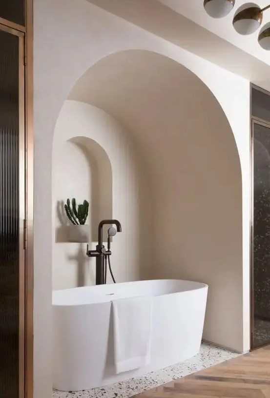 A neutral bathroom with an oval tub placed in a niche, with an additional niche with a potted plant looks jaw dropping