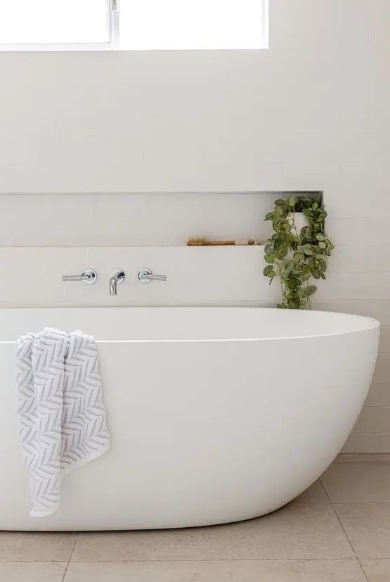 a neutral minimal bathroom clad with white subway tiles, with an oval tub, a niche shelf that is used mostly for decor