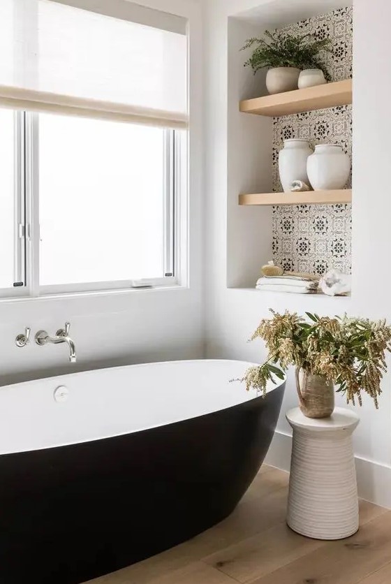 a refined modern bathroom with an oval black tub, a niche with printed tiles with shelves used for decor and storage, greenery in a jug