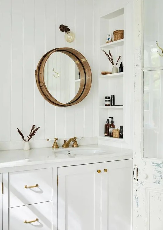 a white farmhouse bathroom clad with skinny tiles, with a large vanity, a niche with shelves to store mostly decor, a round mirror