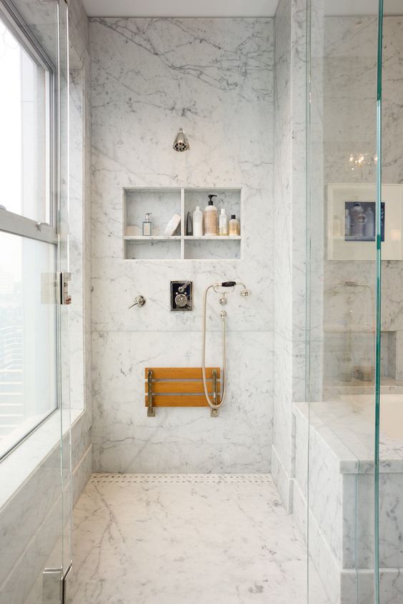 a white marble bathroom with niche shelves used for storage, a folding seat and elegant vintage fixtures