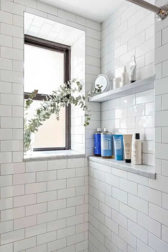 a white shower space with a window, a niche with shelves to store various stuff, greenery is a lovely and airy space