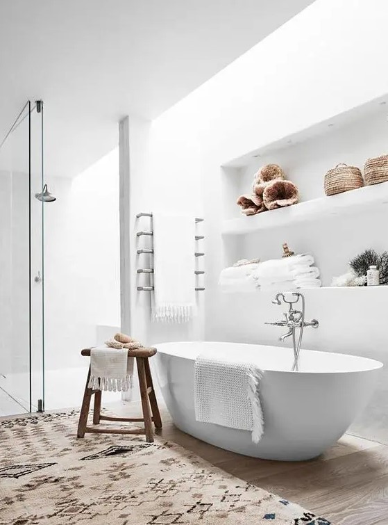 an airy neutral bathroom with a skylight, a shower space, an oval tub, a niche with shelves for decor and storage and a boho rug