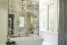 a refined glam bathroom in neutrals with an oval tub, a chic chandelier and a dark mirror wall