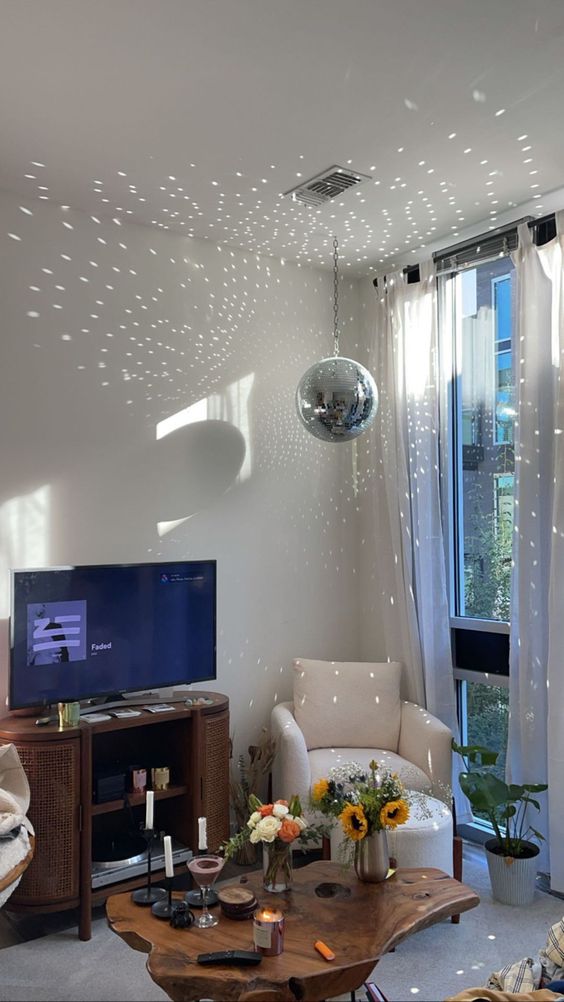 a modern living room with a disco ball at the window, a white chair,a living edge table, a stained TV unit and some plants