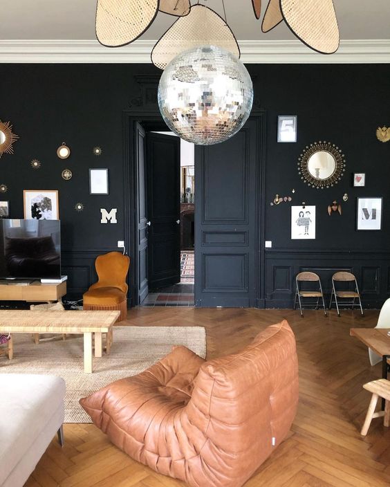 a moody living room with black walls, molding, a woven bench, a rust chair, a brown leather chair, a disco ball and cool gallery walls