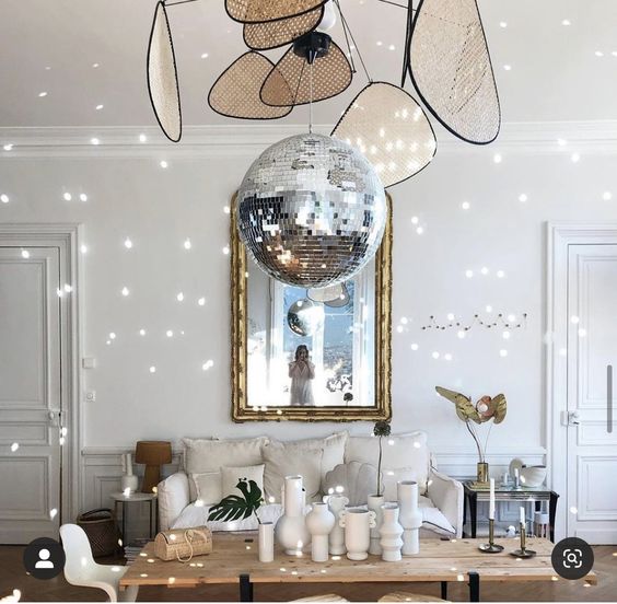 a neutral living room with molding, a neutral sofa, a side table, a table with vases, a large mirror and a disco ball hanging on the chandelier