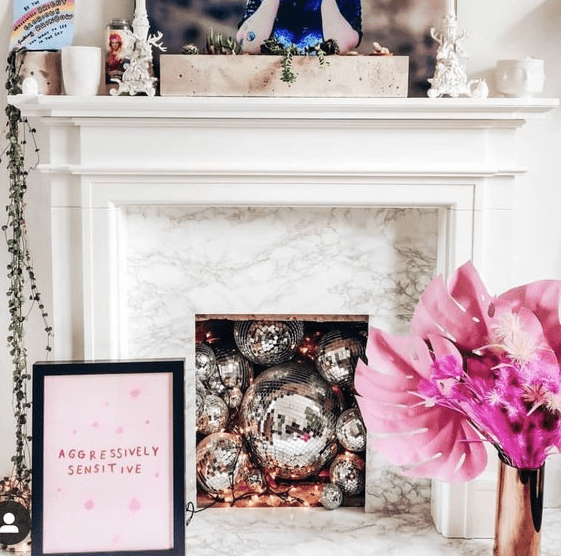 A non working fireplace filled with disco balls, with a mantel with greenery, a vase with pink dried leaves and a sign
