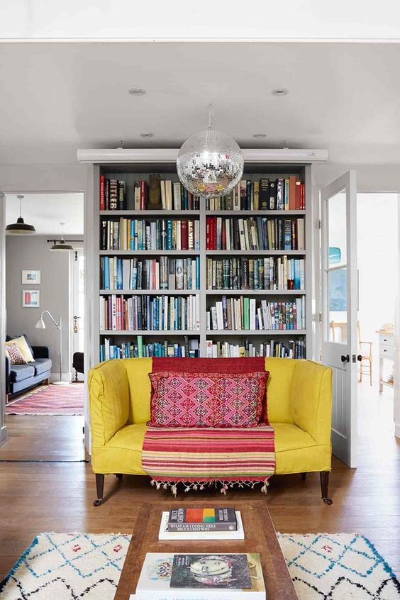 an eclectic and bright space with a yellow loveseat and a blanket, a bold rug, a disco ball and a bookcase behind the loveseat