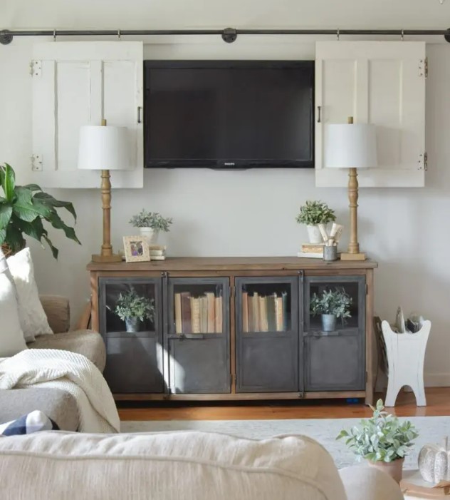 a neutral farmhouse living room with neutral sitting furniture, a TV on the wall hidden behind white barn doors and a metal and wood storage unit