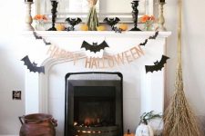 a Halloween mantel with bats, candles, pumpkins, orange blooms, a wheat bundle and some pumpkins and a broom on the floor