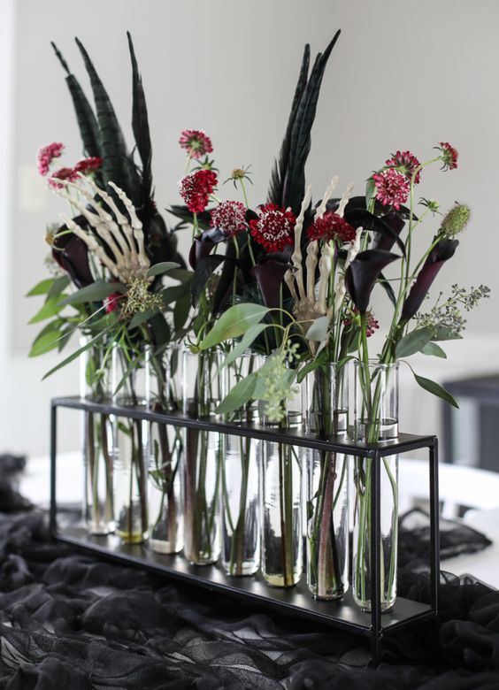 a very elegant Halloween centerpiece of a stand with test tubes, deep purple callas and burgundy blooms, greenery, feathers and skeleton hands