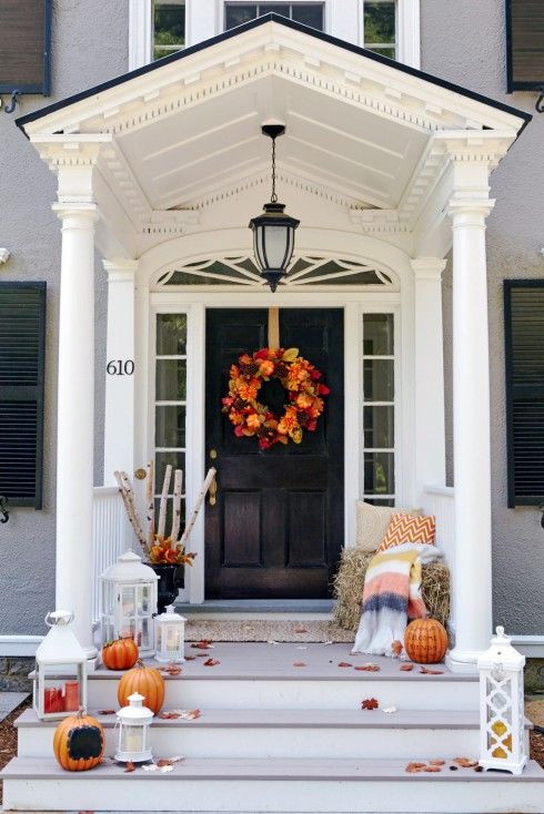 a Thanksgiving porch with white lanterns, orange pumpkins and candles, hay and branches and a fall leaf wreath