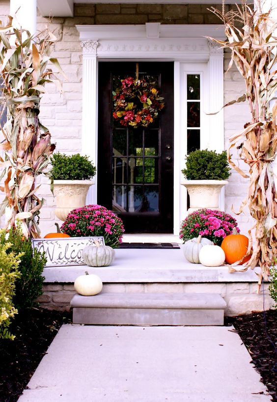 a bright and chic fall porch with corn husks, pink blooms in pots, natural pumpkins and a fall wreath