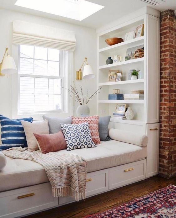 a cool windowsill reading nook with bright pillows, wall sconces and built in shelves and neutral shades