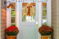 a cozy and colorful Thanksgiving porch with potted blooms, potted greenery, a corn husk wreath and stacked pumpkins with lights