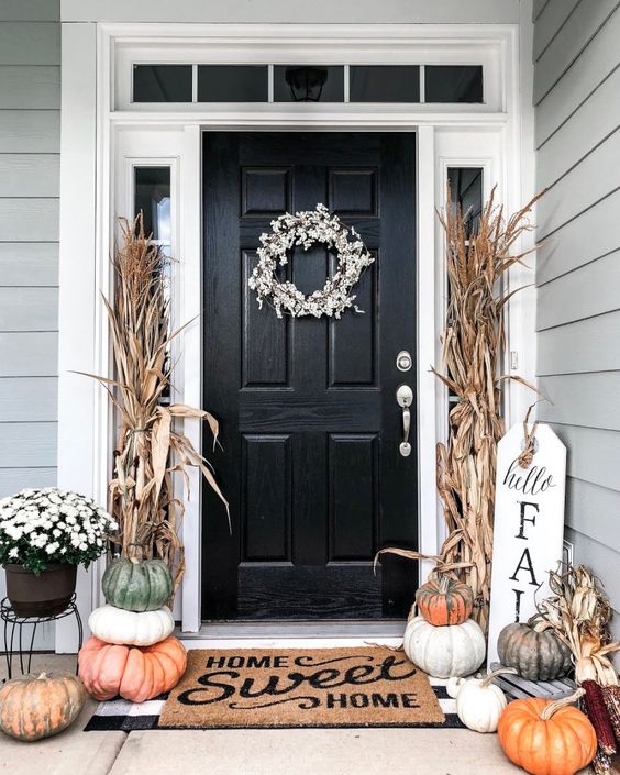 a stylish and casual fall porch with stacked heirloom pumpkins, corn husks, a floral wreath and potted blooms