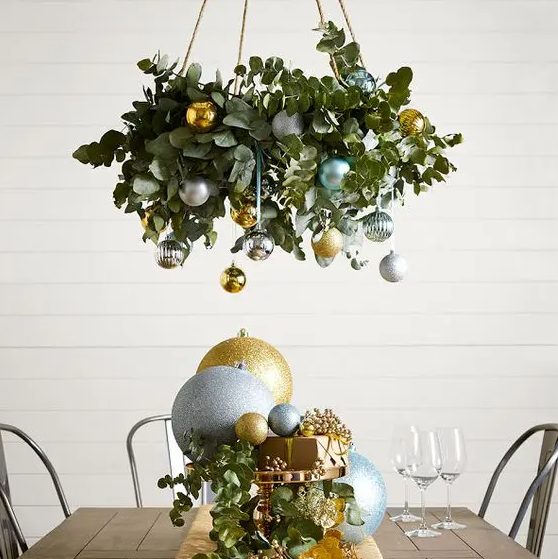a chic holiday chandelier of eucalyptus, silver, gold and green ornaments and matching oversized ones on the table