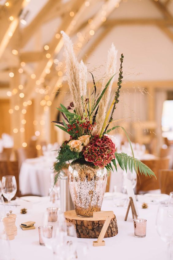 a chic wedding centerpiece of a mercury glass vase and rust and red blooms, pampas grass and ferns