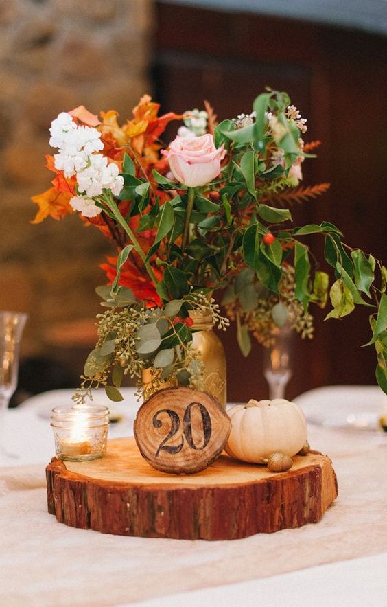 a fall wedding centerpiece of a wood slice, pumpkins, candles and bright fall blooms in a gold jar
