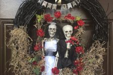 a black vine wreath with hay, blooms, a bunting and skeletons dressed in a chic way will decorate your front door for Halloween