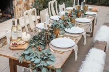 a chic outdoor fall tablescape with a greenery and berry runner, candles, amber goblets, white pumpkins and gold cutlery
