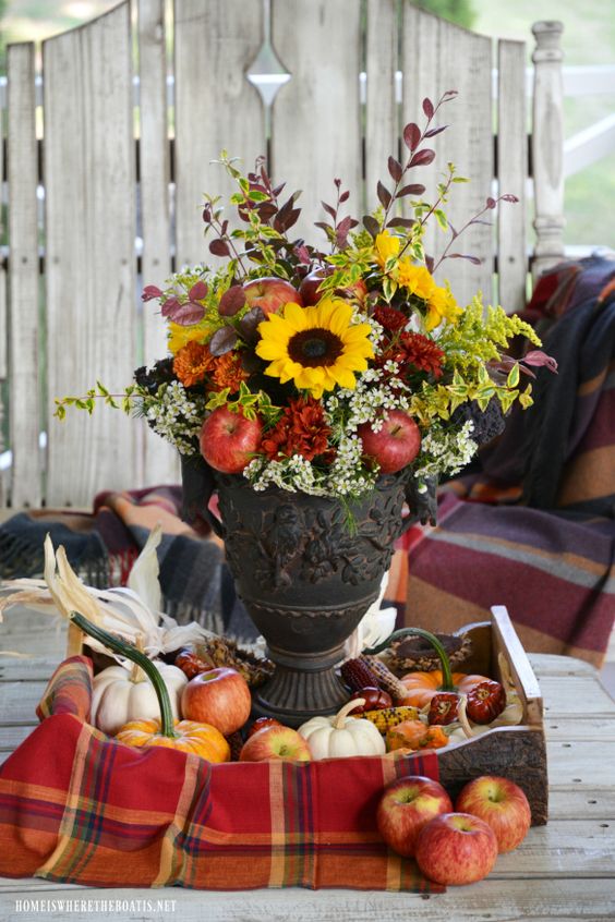 a bright fall arrangement with bold blooms, greenery, red apples and dark foliage placed in a tray with apples, gourds and pumpkins