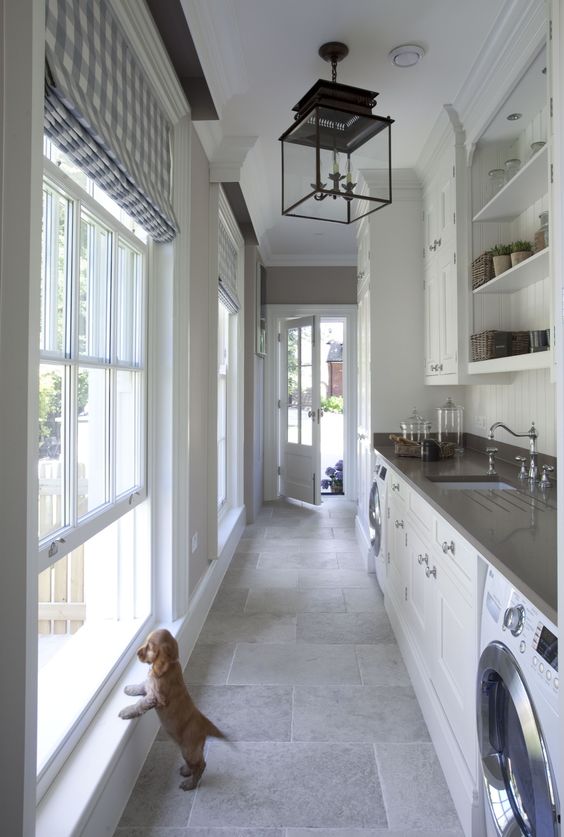 a long and narrow white mudroom and laundry with shaker cabinets, open shelves, built-in appliances and a couple of large windows