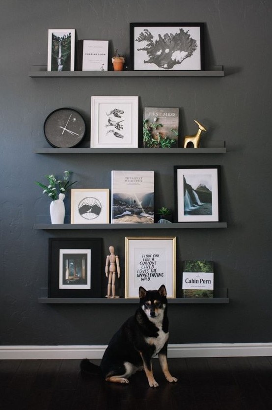 a black wall with black ledges and chic artworks, books, statuettes of various kinds, a clock and some potted greenery