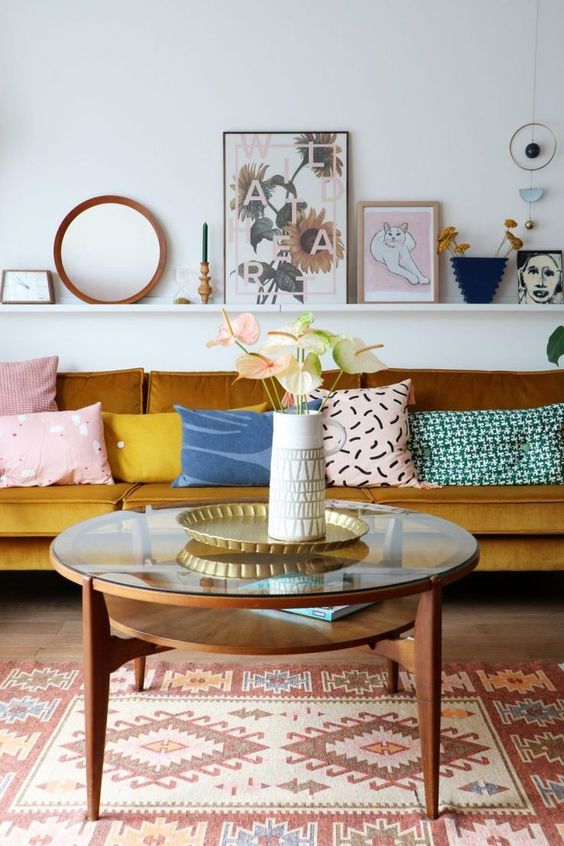 a bright living room with a mustard sofa, a round tiered coffee table, a boho rug, a ledge with artwork and a mirror