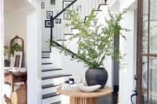 a chic and stylish entryway with a black and white gallery wall over the stairs, a wooden table with a large planter and greenery and a stool