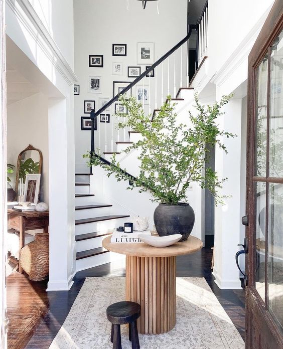 a chic and stylish entryway with a black and white gallery wall over the stairs, a wooden table with a large planter and greenery and a stool