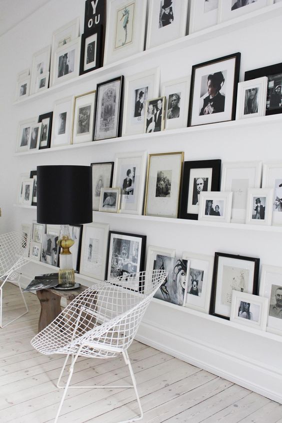 a floor to ceiling gallery wall with black and white artwork, white metal chairs, a wooden bench and a cool lamp