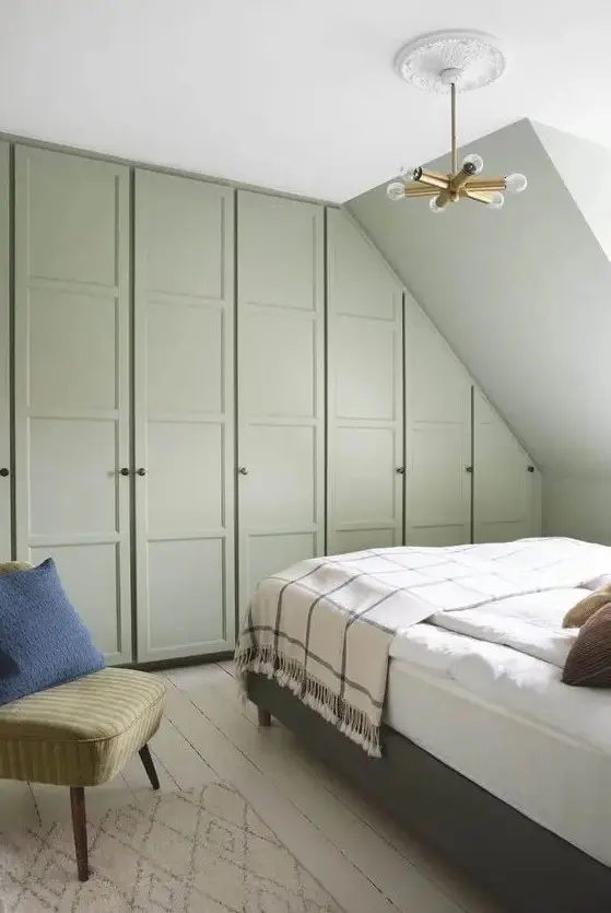 a green bedroom with built-in attic and usual wardrobes, a grey bed with neutral bedding, a green chair and a rug plus an elegant chandelier