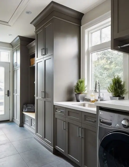 a grey farmhouse laundry mudroom with shaker cabinets, a built-in rack and bench and some appliances next to the sink