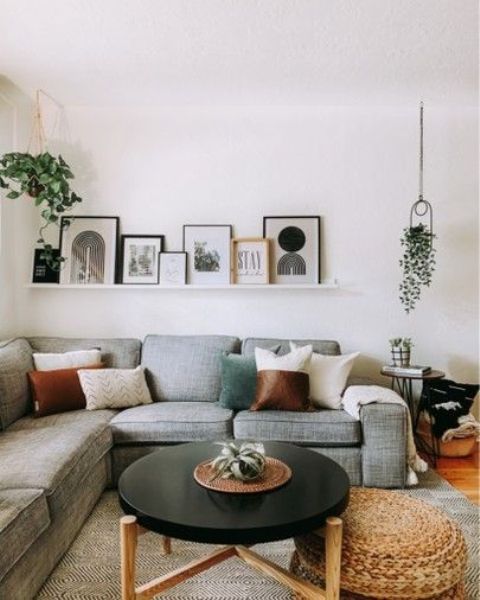 a modern living room with a grey sectional and lots of pillows, a ledge with some art, a black coffee table and some greenery