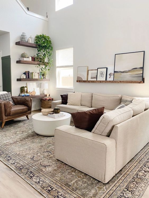 a modern living room with a large neutral sectional, a stained ledge with art, a white coffee table, a brown leather chair and some stained shelves