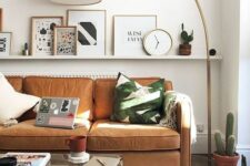 a modern living room with an amber leather sofa, a ledge gallery wall with potted plants, a clock and some art, a glass coffee table