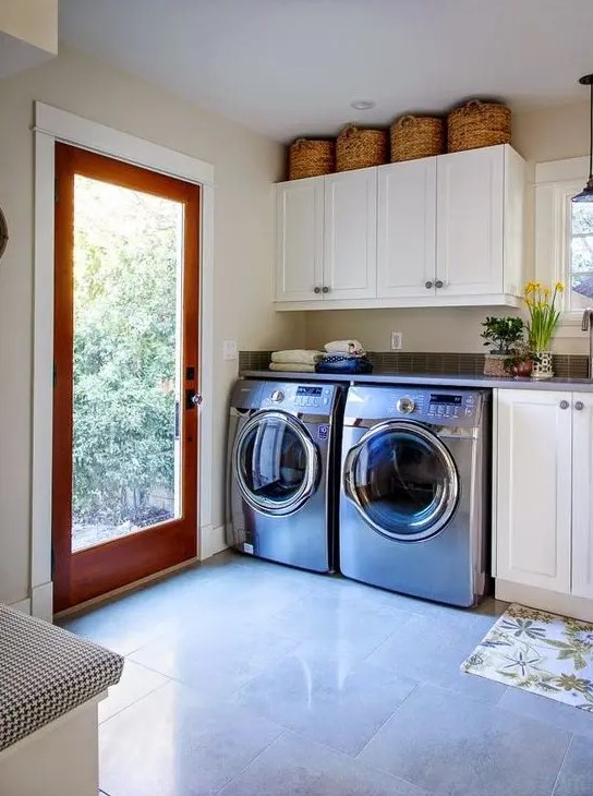 a neutral spacious mudroom laundry with white shaker cabinets, an upholstered bench, a washing machine and a dryer and a glass door