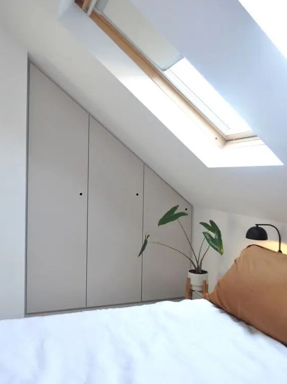 a small attic bedroom with a built-in storage unit with grey doors, a bed, a black lamp and a potted plant plus some skylights
