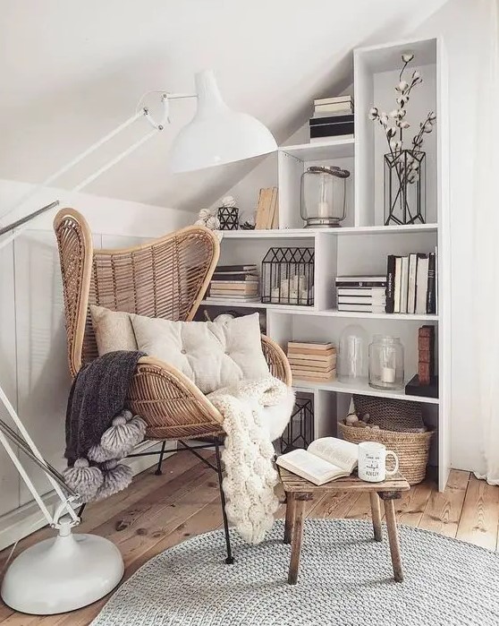 a small attic reading nook with a white box shelving unit, a rattan chair and a wooden stool, a floor lamp and some textiles