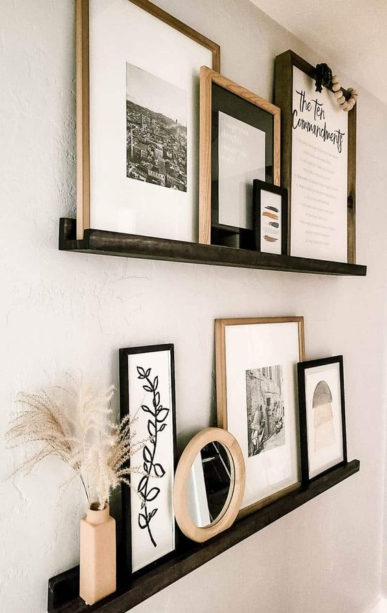 a stylish gallery wall with black ledges, artwork, photos and signs, some dried grasses and a mirror in a wooden frame